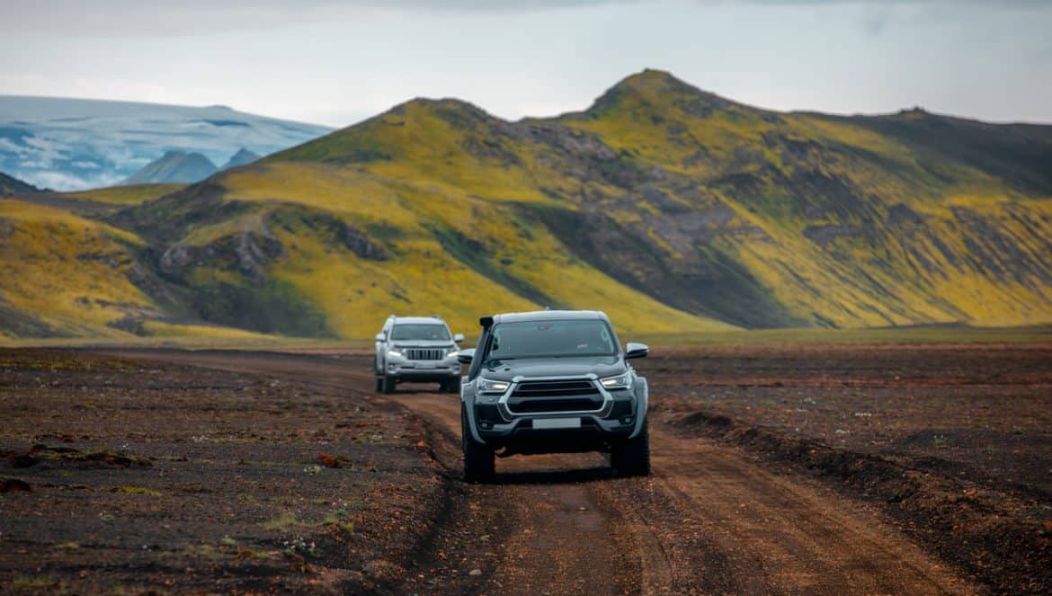 Two off-road vehicles driving along a rugged dirt road through the volcanic terrain of Iceland. The lush green moss covering the mountains contrasts sharply against the dark soil and rocky path. The scene reflects the adventurous spirit of Icelandic exploration, with glaciers visible in the background