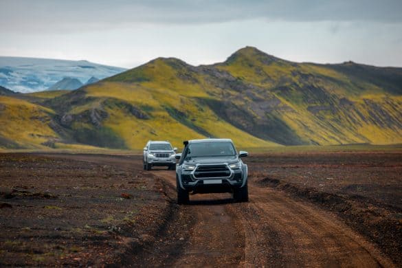 Two off-road vehicles driving along a rugged dirt road through the volcanic terrain of Iceland. The lush green moss covering the mountains contrasts sharply against the dark soil and rocky path. The scene reflects the adventurous spirit of Icelandic exploration, with glaciers visible in the background