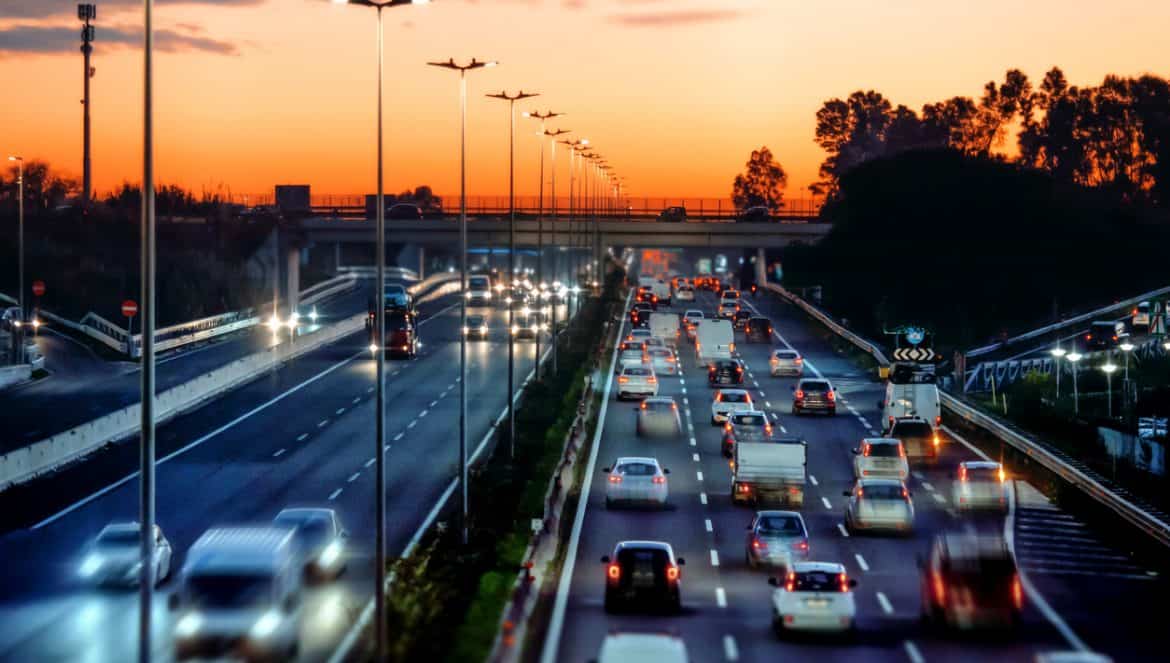 Busy highway with plenty of cars and trucks lined up with lights on at sunset. City life with traffic jams. Motion blur, warm sky.
