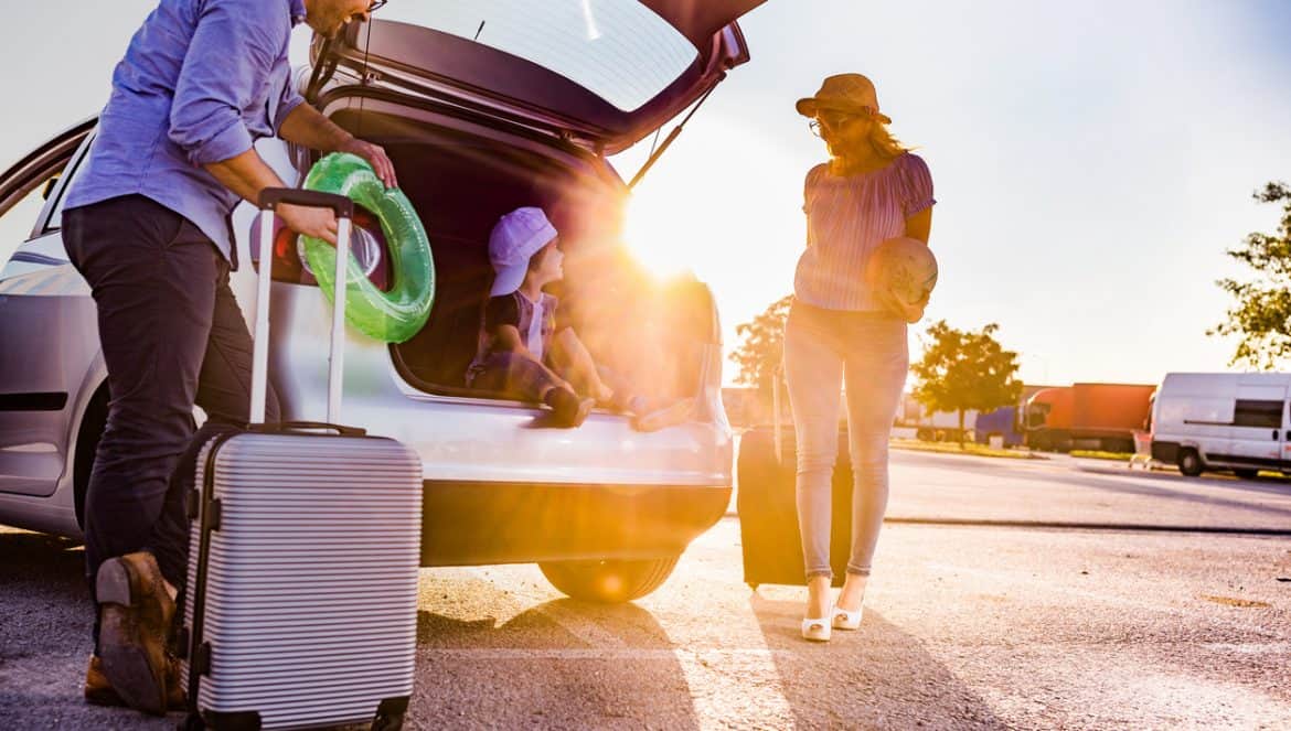 Cute little boy sitting inside of a car trunk ready for summer vacation while his parents are talking to him.
