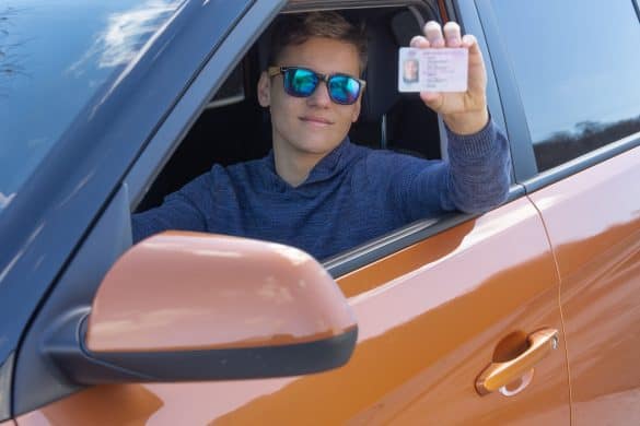 Happy teenager is showing his driving license from open car window. Driving school concept