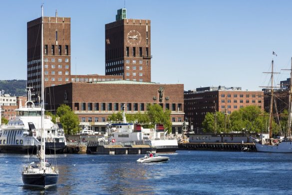 A sail boat sails from the marina in the Oslo fjord in front of the City hall building in Norway capital city on a sunny day.