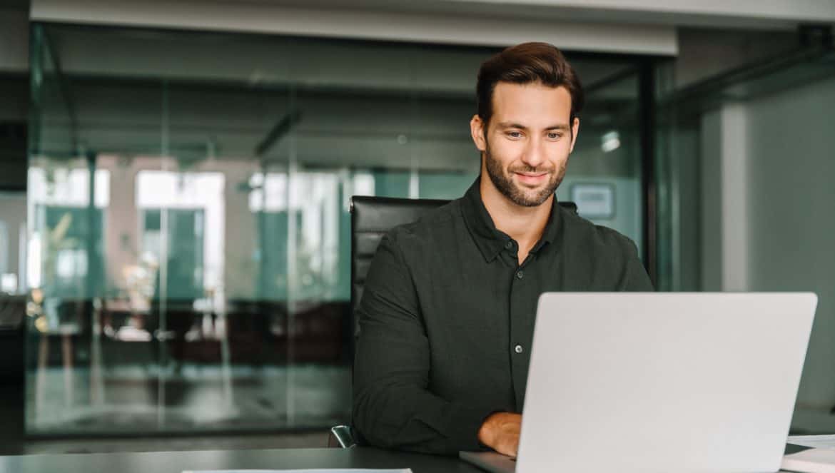 Young professional it specialist European business man working on laptop pc sitting at desk in modern office. Middle-aged latino businessman using computer technology app for work online. Copy space